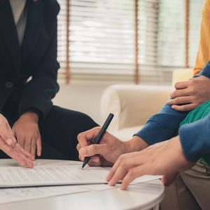 Happy young Asian couple and realtor agent. Cheerful young man s Happy young Asian couple and realtor agent. Cheerful young man signing some documents while sitting at desk together with his wife. Buying new house real estate. Signing good condition contract.